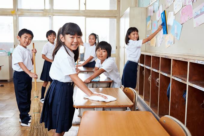 Japanese elementary school students smiling while actively cleaning their classroom desks and floor during Souji time